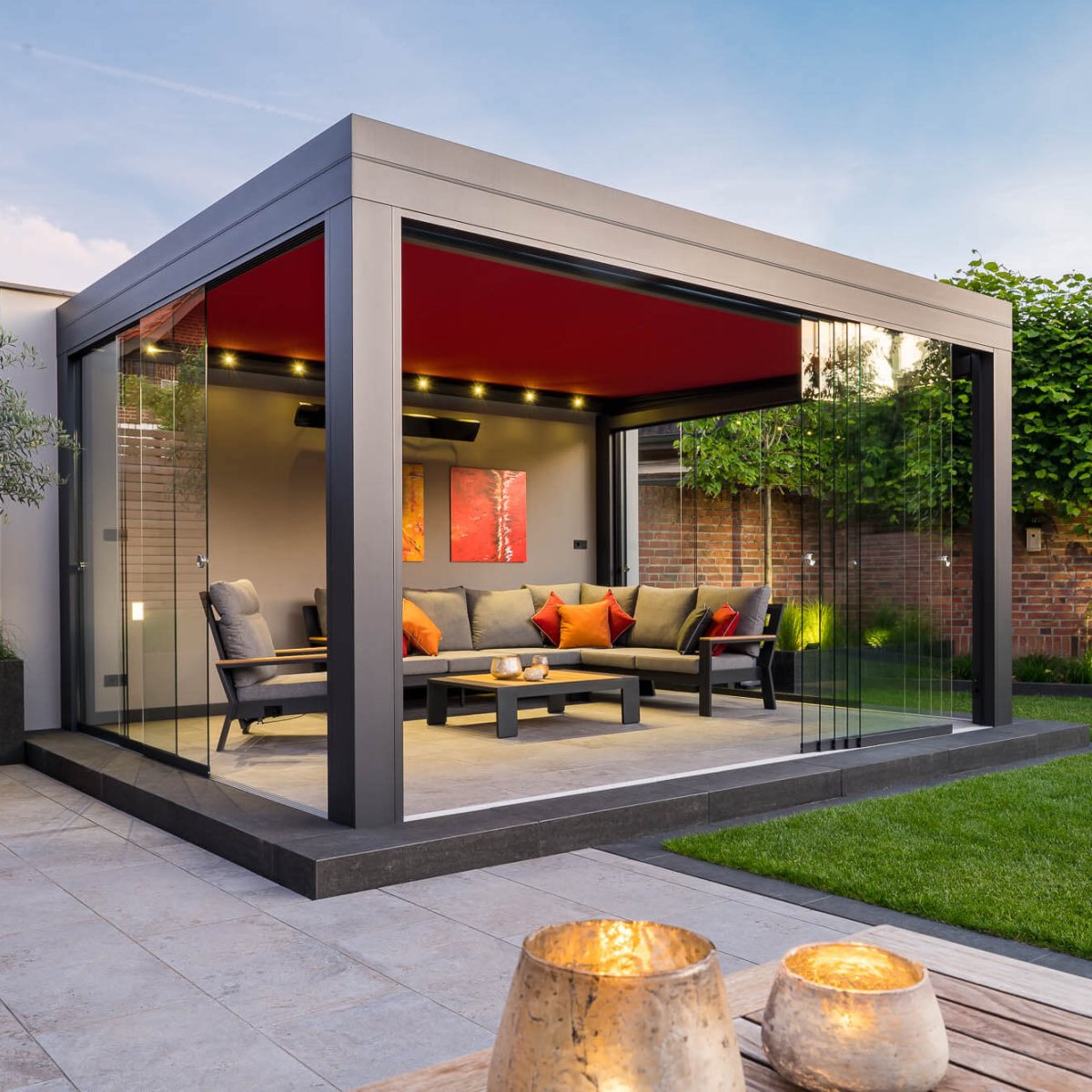 Moderner verglaster Gartenpavillon mit Sitzgruppe und roter Decke im gepflegten Garten