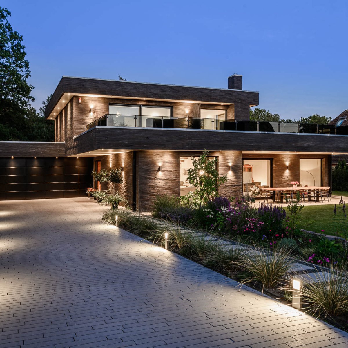 Modernes zweigeschossiges Haus mit beleuchtetem Eingang, Einfahrt und Garten bei Dämmerung