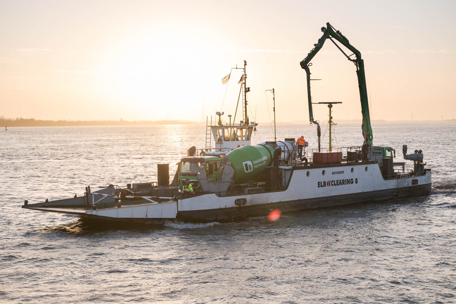 Arbeitsfähre mit Betonmischer und ausgeklappter Betonpumpe fährt bei Sonnenuntergang über breiten Fluss; Crew an Deck.