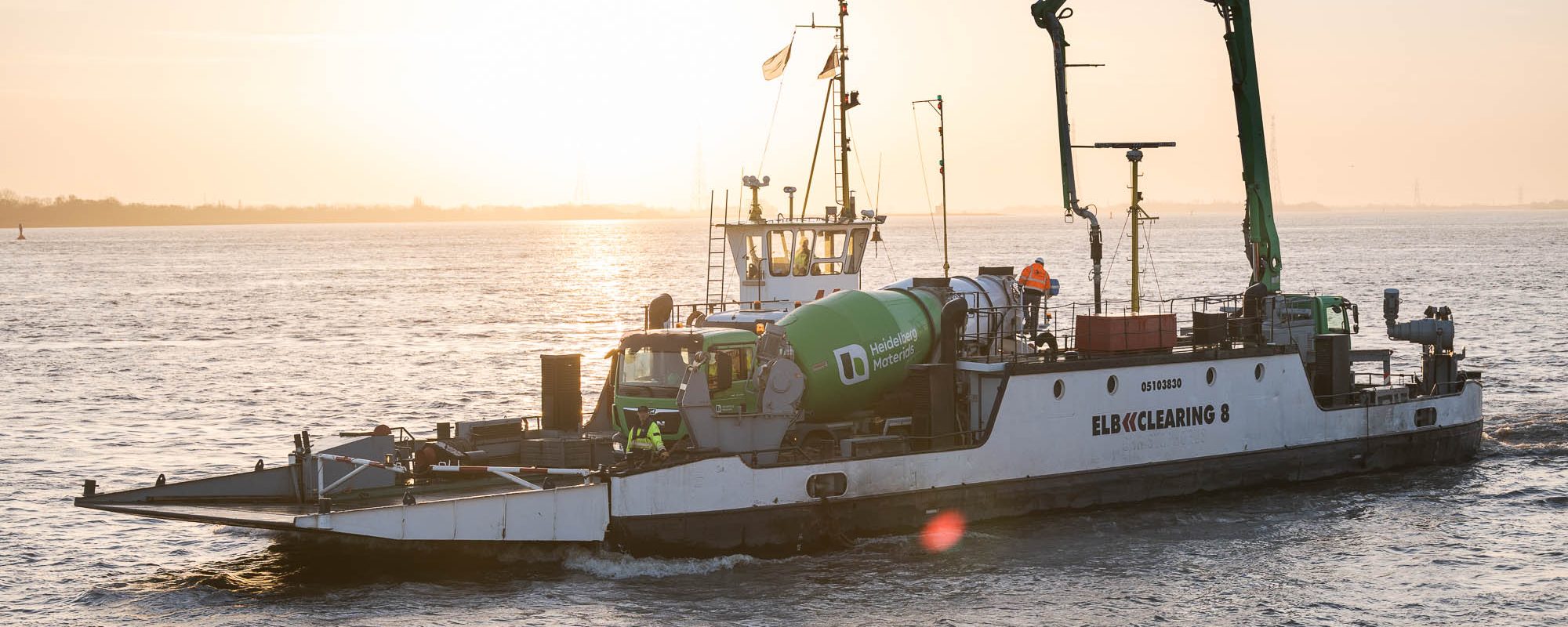 Arbeitsfähre mit Betonmischer und ausgeklappter Betonpumpe fährt bei Sonnenuntergang über breiten Fluss; Crew an Deck.