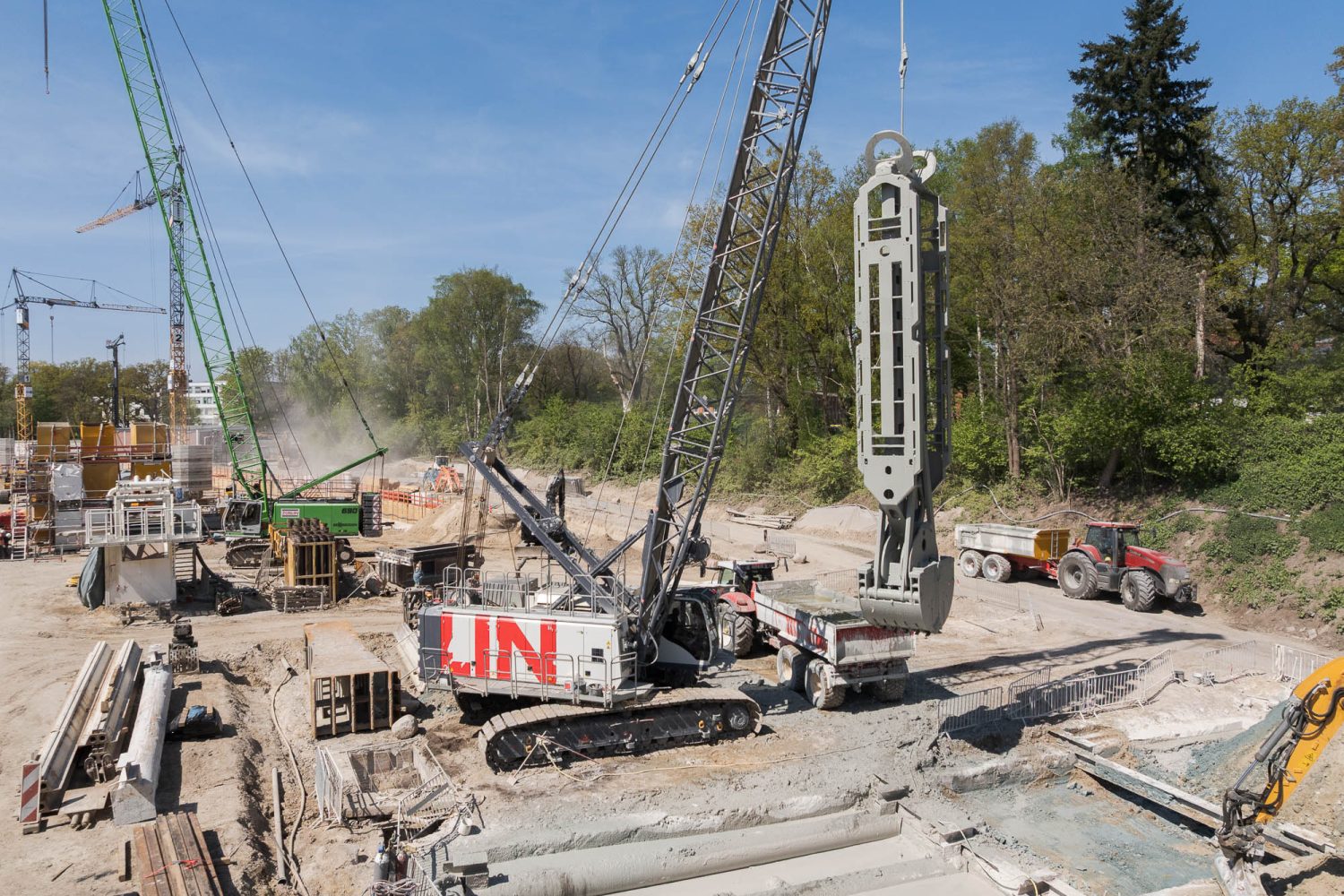 Raupenkran mit Greifer belädt Kipp-Lkw mit Aushub; Tiefbauarbeiten auf Großbaustelle mit weiteren Baumaschinen.